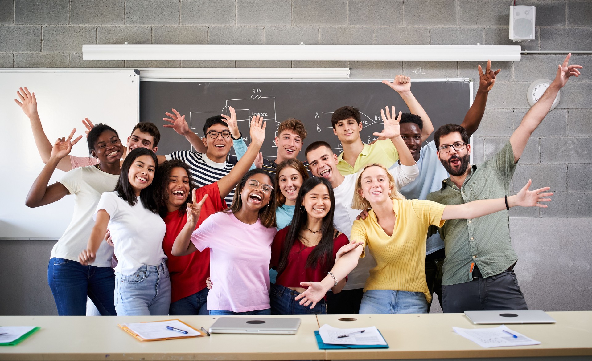Portrait of a cheerful group of students celebrating in class looking at the camera. Happy Young college people of different ethnicities posing for a photo in the classroom hands up